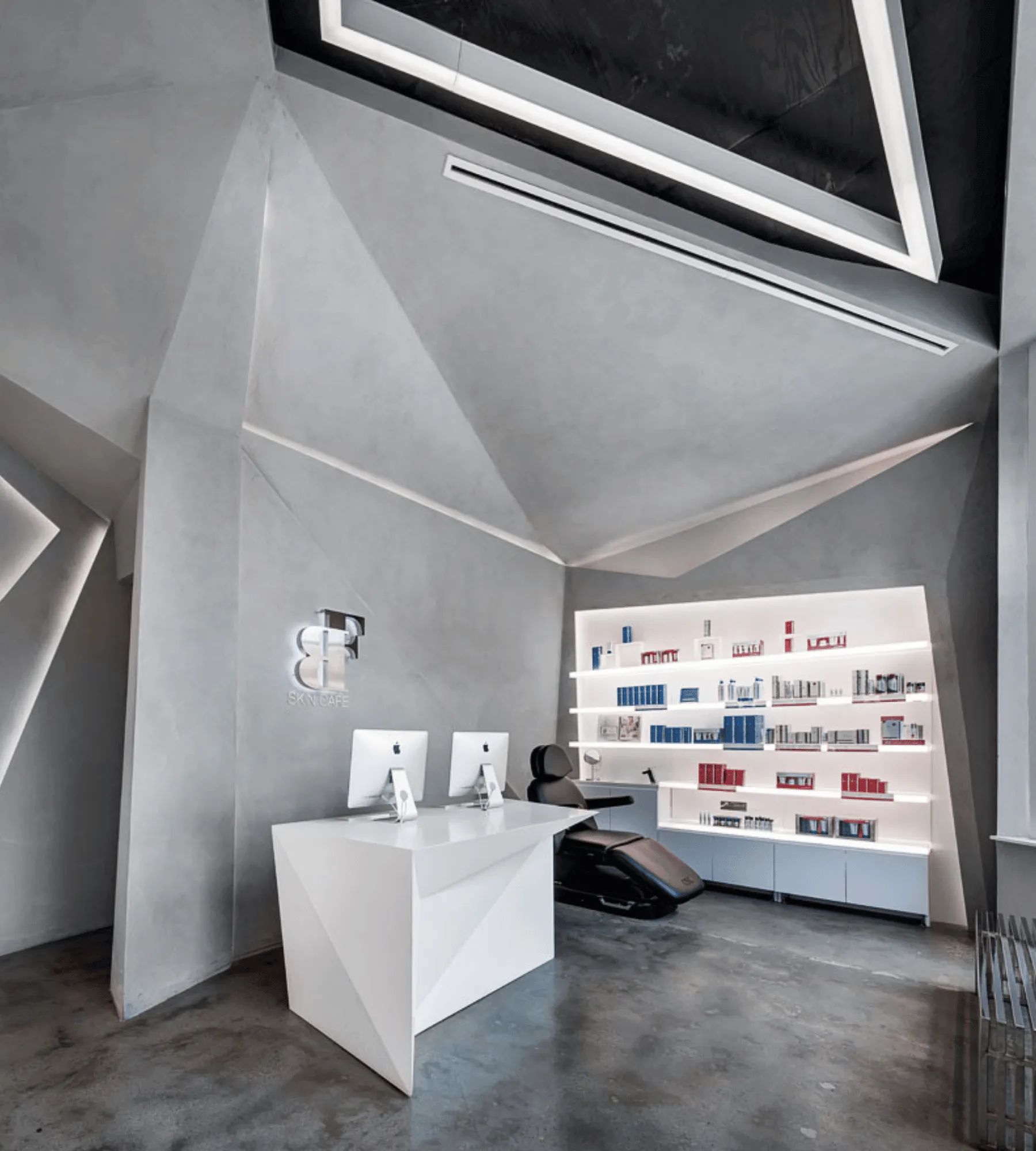 The reception room: a faceted white desk with two screens, the chrome logo on the concrete wall and a backlit product wall behind.