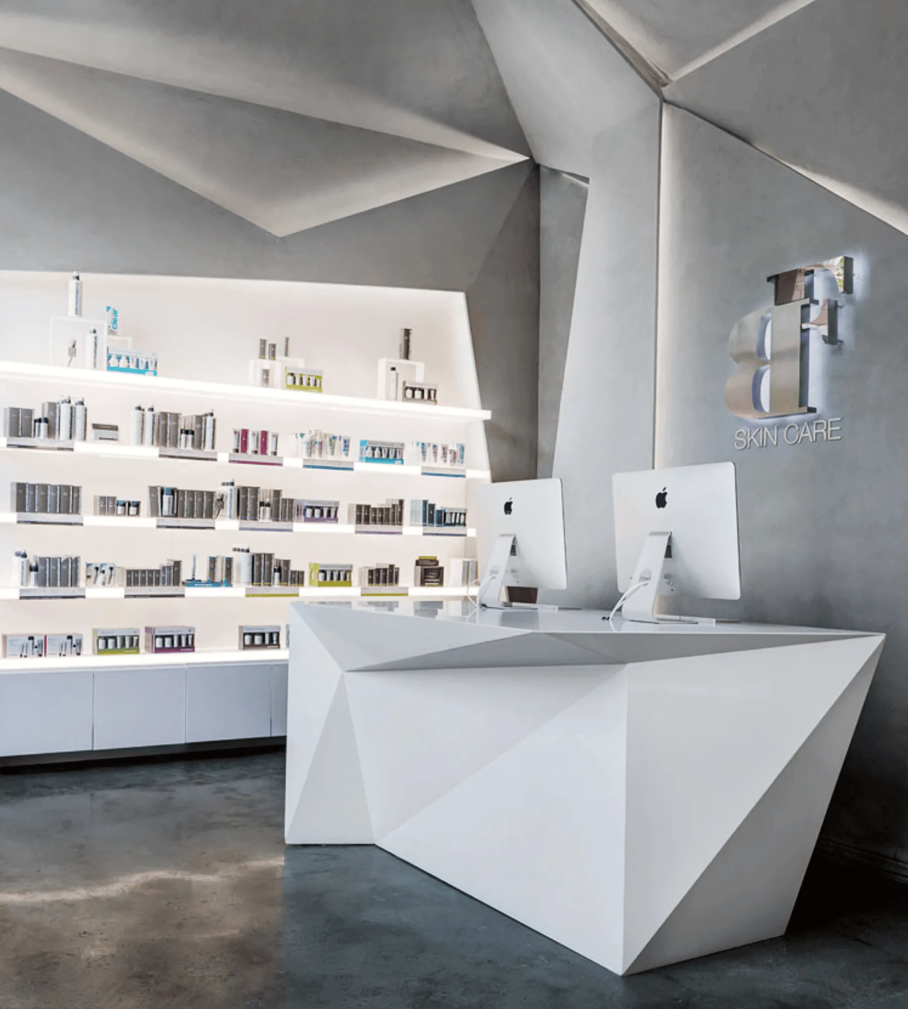 A backlit shelf wall of skin care product beside the white faceted reception desk, under folded concrete ceiling planes.