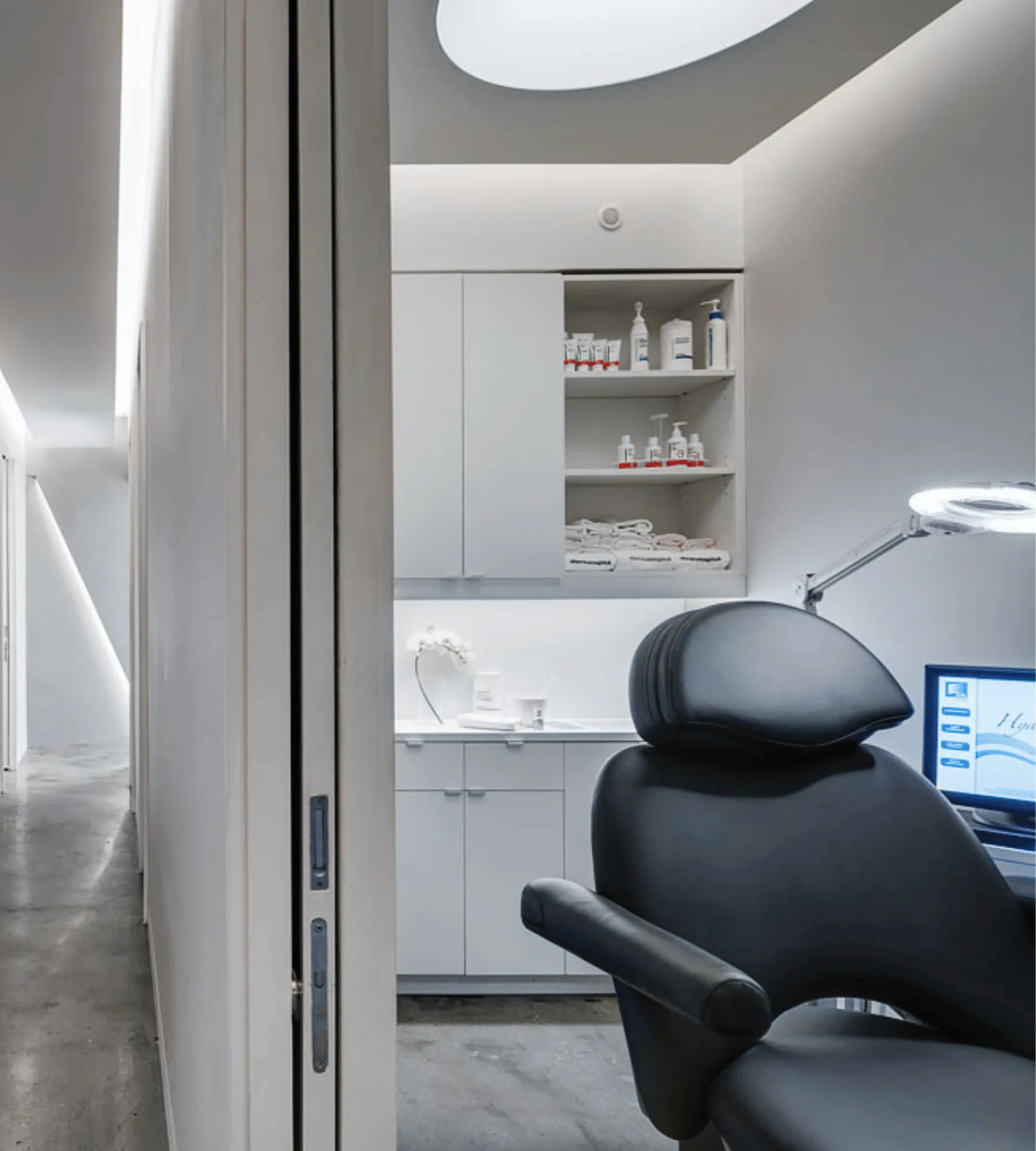 A treatment room seen through its sliding door: a black reclining chair under a magnifier lamp, shelves of product and folded towels, an oval skylight overhead.