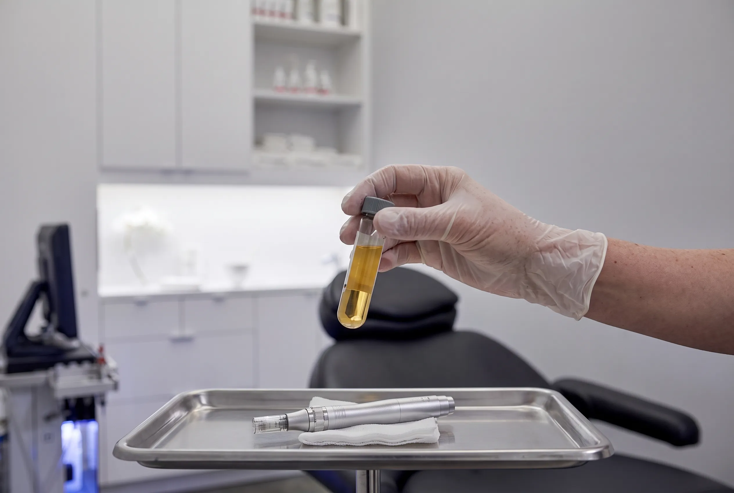 A gloved hand holding a sealed vial of golden serum above a steel tray laid with a pen and gauze.