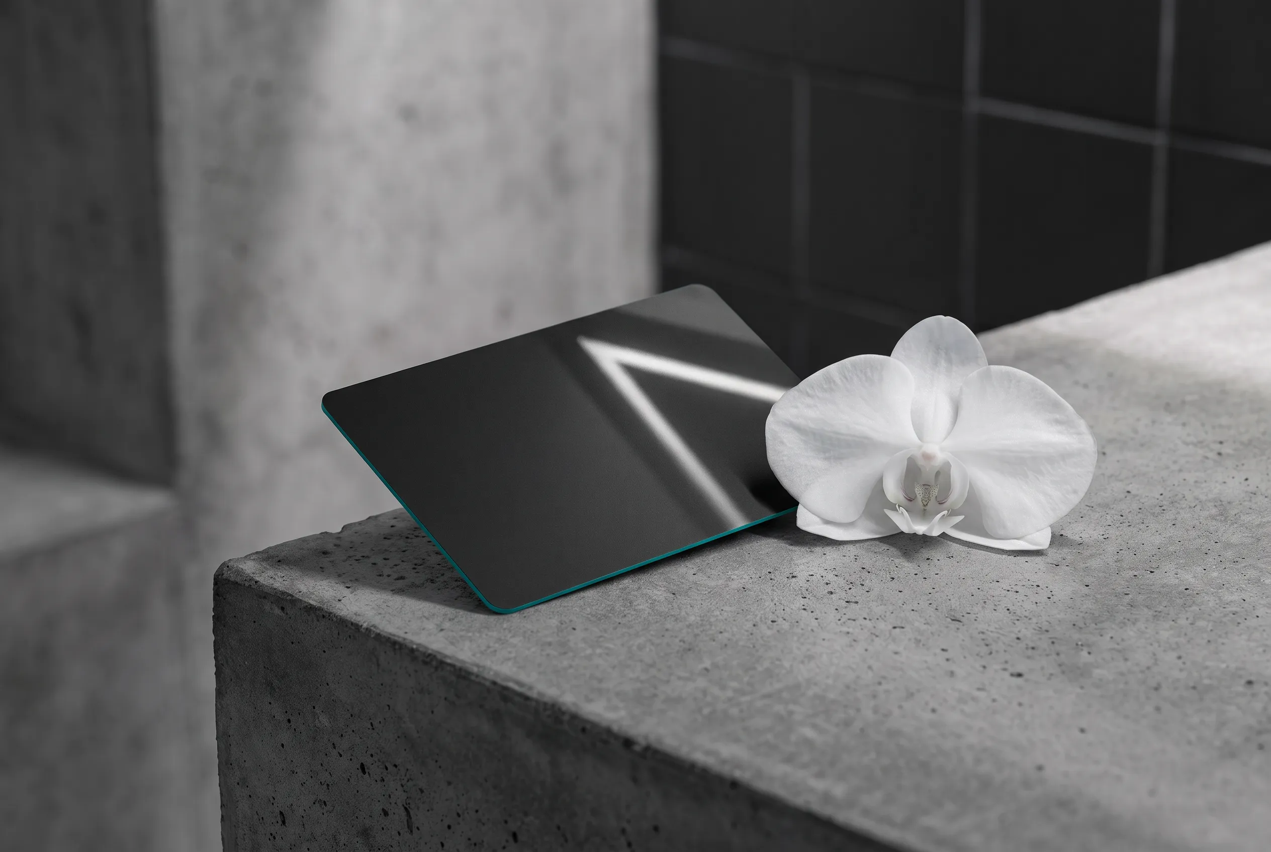 A matte black gift card with a painted teal edge lying on raw concrete, the ceiling light line reflected along its length.